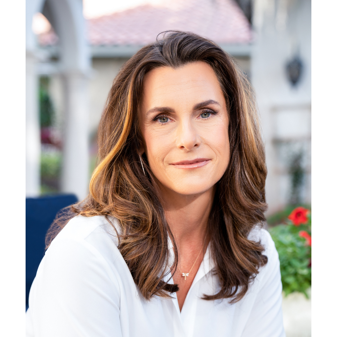 Close-up of a woman with wavy brown hair and blue eyes, wearing a white top and a small cross necklace, outdoors in a garden setting.
