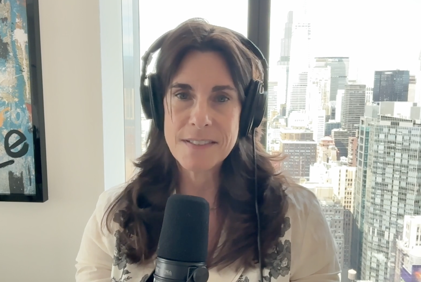 A woman with brown hair wearing headphones speaking into a microphone, in front of a window with a cityscape view of tall buildings.