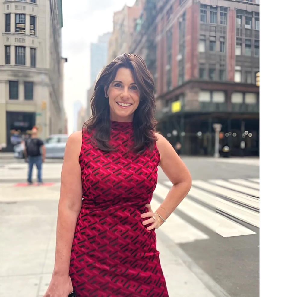 A woman with shoulder-length dark brown hair in a red sleeveless dress with a black pattern, standing on a city street with tall buildings in the background, smiling at the camera.