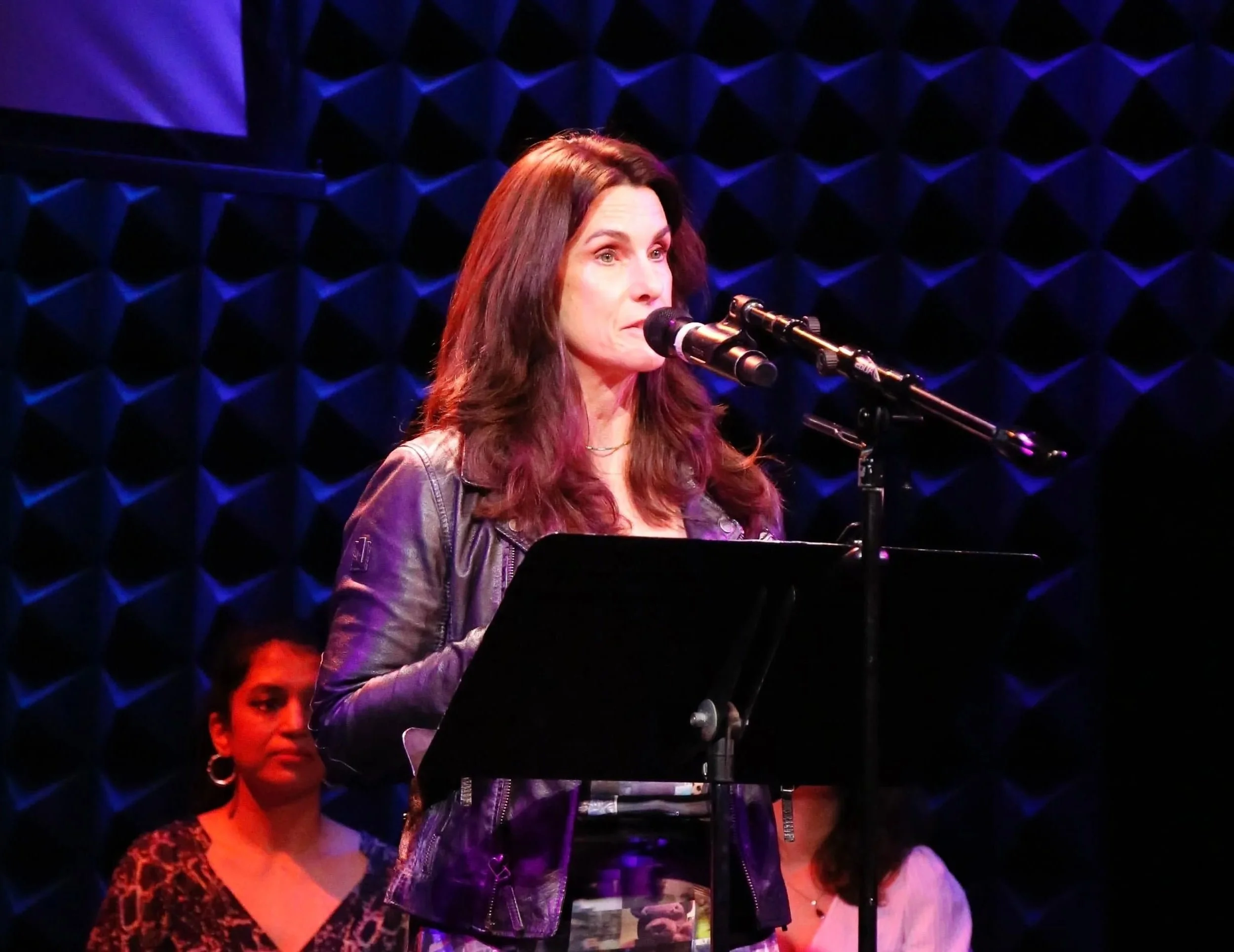 A woman with long dark hair and a leather jacket standing at a microphone on stage, with a woman in a leopard print top visible in the background.