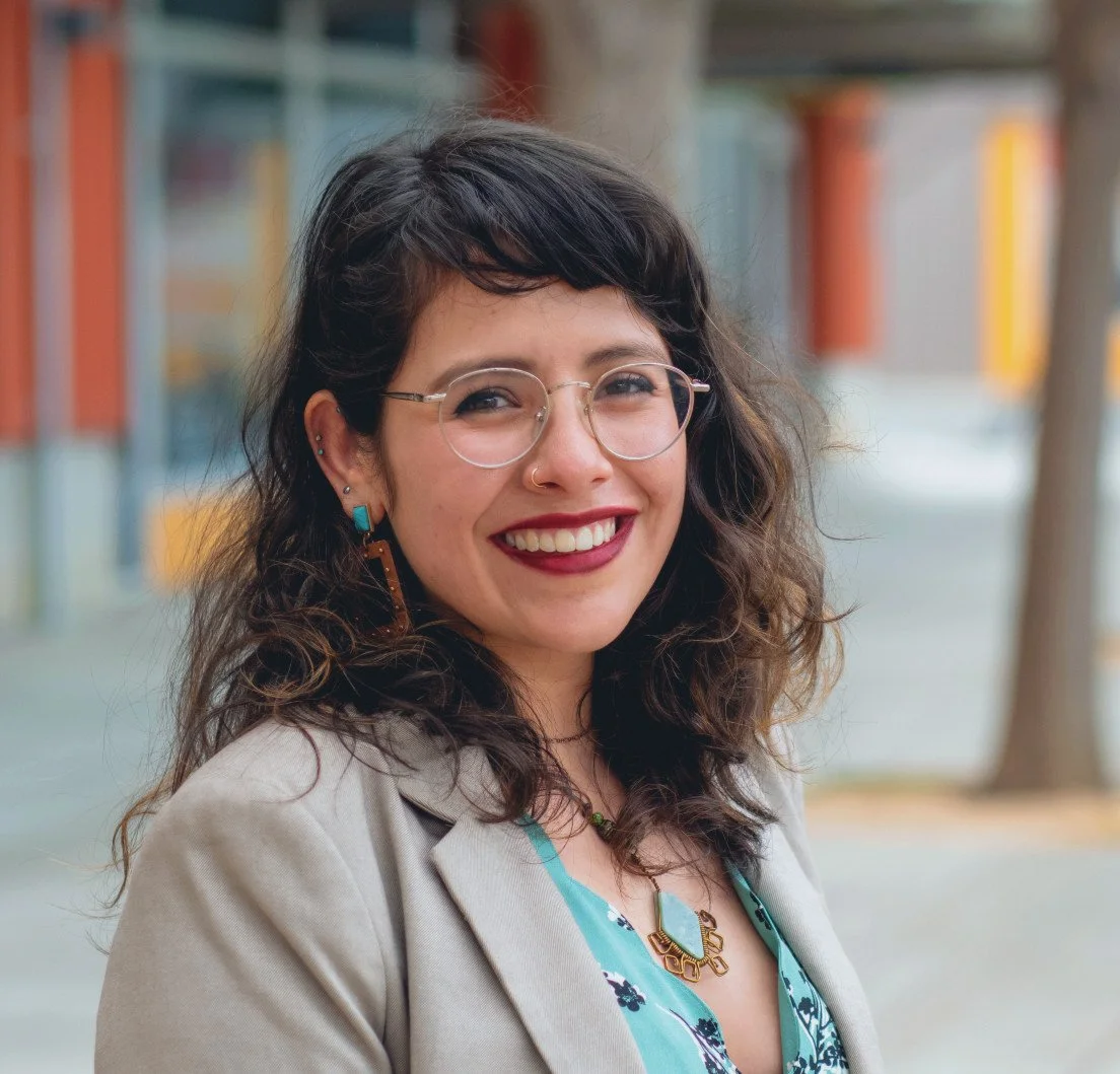 A woman with black, wavy hair, glasses, and a bright smile, standing outdoors in front of a colorful building.