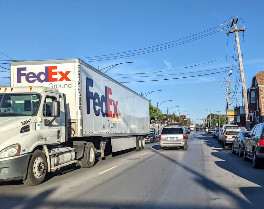 A large FedEx freight truck dominates a narrow urban street, squeezing past parked cars in a residential neighborhood