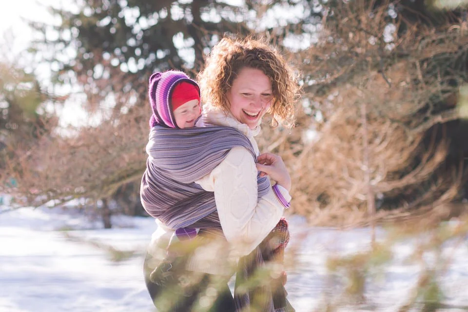 Mom and baby smiling outside in the snow near sunset at the Arboretum in Ottawa. Mom is wearing a white sweater and baby is wrapped in a purple wrap and is wearing a red hat.