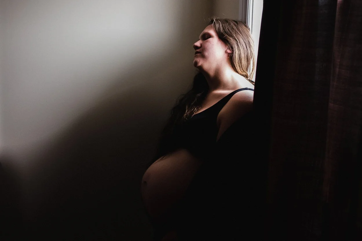 Woman in labour standing at a window in a slit of light. The rest of the scene is dark.