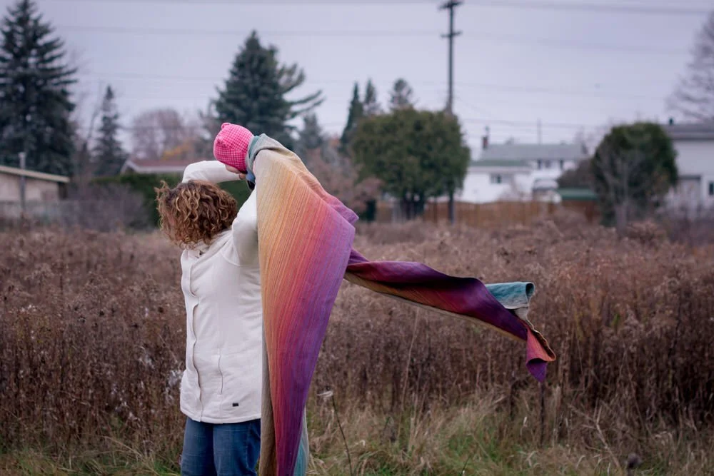 Mom holding baby above her back about to put baby on her back. The wrap is blowing in the wind and baby is laughing.