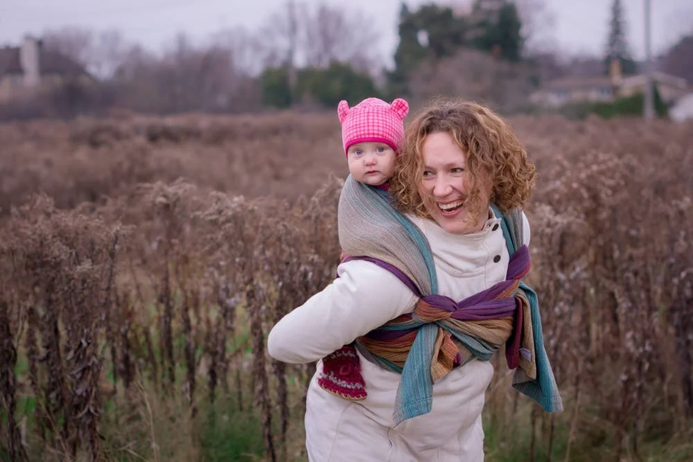 Mom and baby at Kilborn Allotment Garden wearing a colourful baby carrier wrap and baby is in a pink hat. They are surrounded by brown coloured plants.