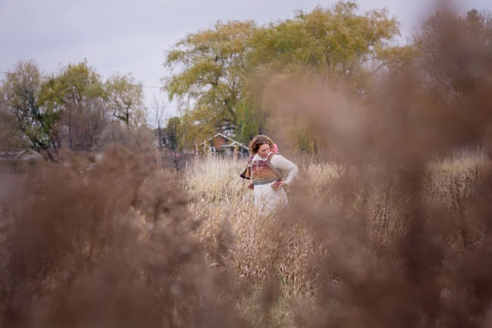 Mom and baby walking in tall grass with colourful wrap and large weeping willows behind them in the fall.