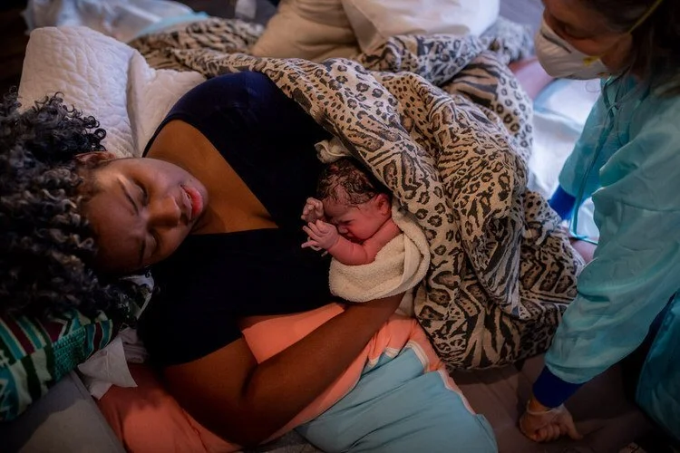 Mom holding her newborn son right after birth. He is wrapped in towels and blankets as midwife overlooks.