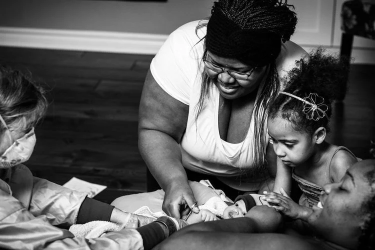 Big sister watches as her aunt cuts her newborn brother's cord while midwife guides. Mom looks radiant and happy.
