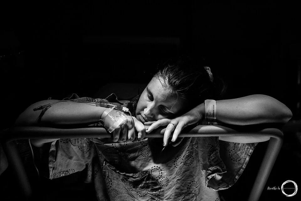 Woman in labour at Montfort Hospital during birth. She is in the dark under one light resting on a bar with an iv in her hand. She looks peaceful.