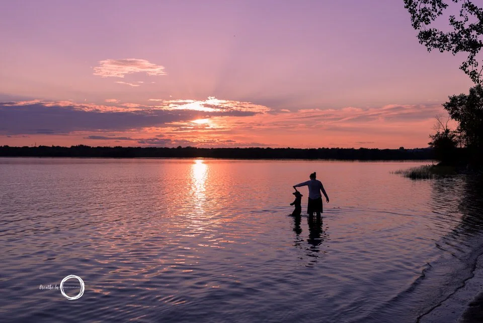 Mom and daughter in Ottawa River at sunset at Westboro Beach. The sky is pink and orange and the sun is reflected in the water.