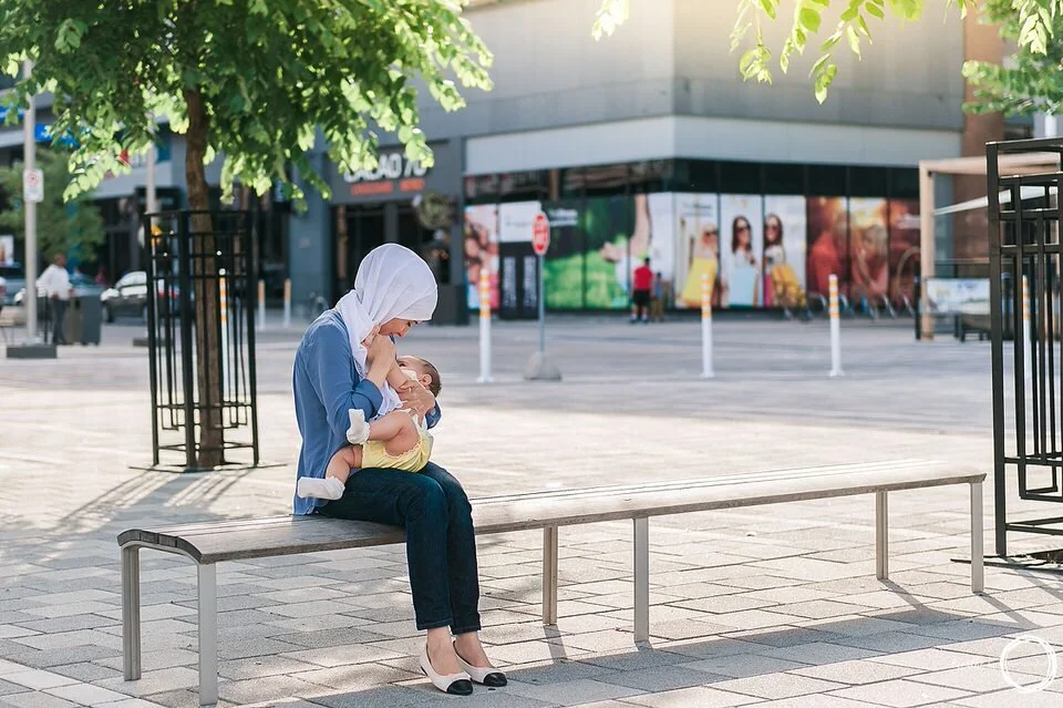 Woman sitting on a bench with her baby breastfeeding near a tree with the sun setting behind her surrounded by stores in the background at Landsdowne Park, in Ottawa.