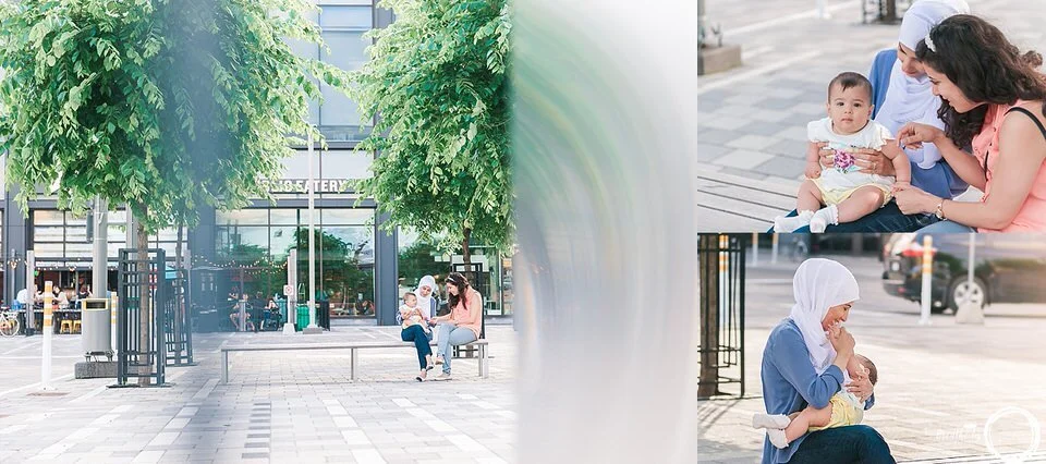 Mom, baby and friend are sitting together on a bench between two large green trees at Landsdowne Park, they are smiling and laughing together.