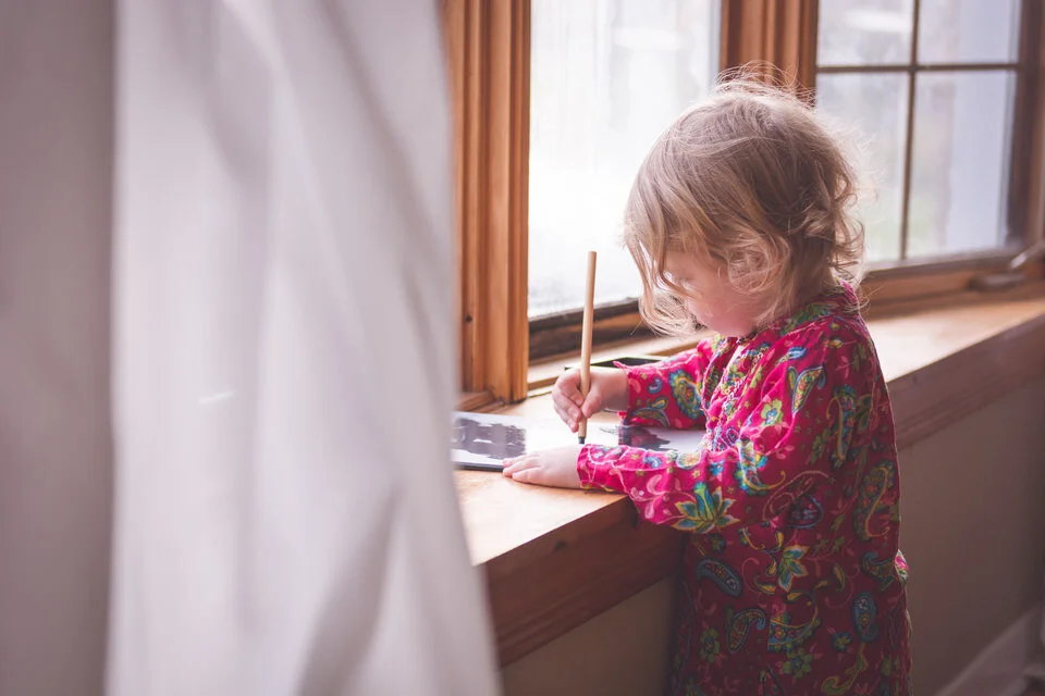 Toddler standing in window light at her home painting with water. She is wearing a colourful pink shirt and has soft curly hair.