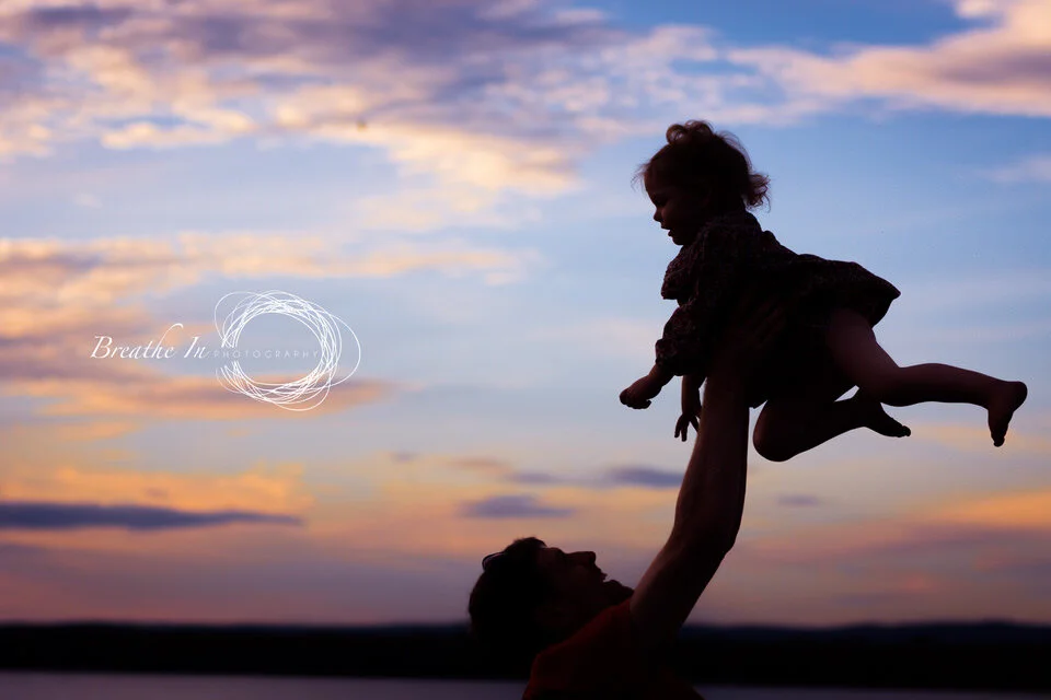 Dad lifting his toddler up above him at sunset at Britannia Beach in Ottawa. The sky behind them is pink, orange and blue. They are in shadow.