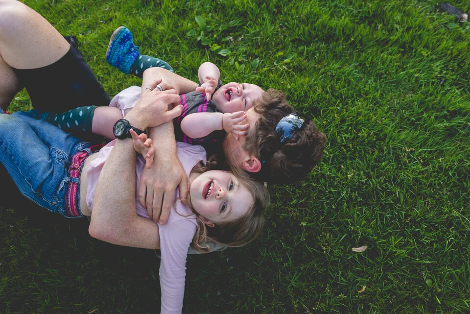 Dad holding his two children. One is a baby and the other is 4 years old. They are laying in the grass laughing and giggling.