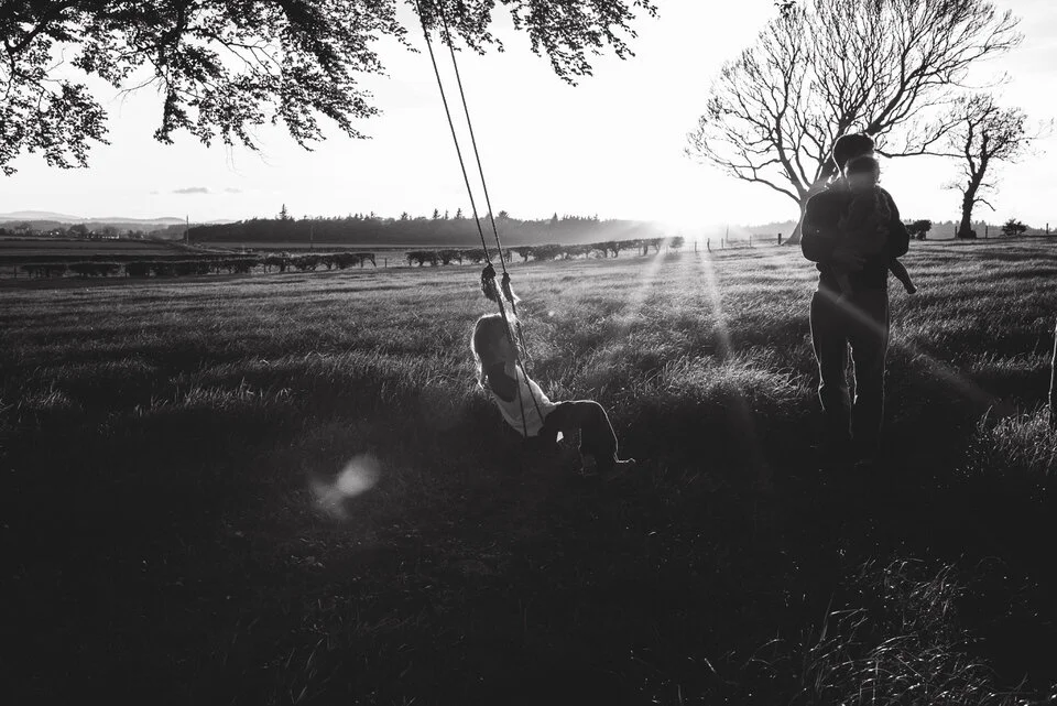 Dad standing holding a baby while bigger child is swinging on a swing at sunset in an open field.