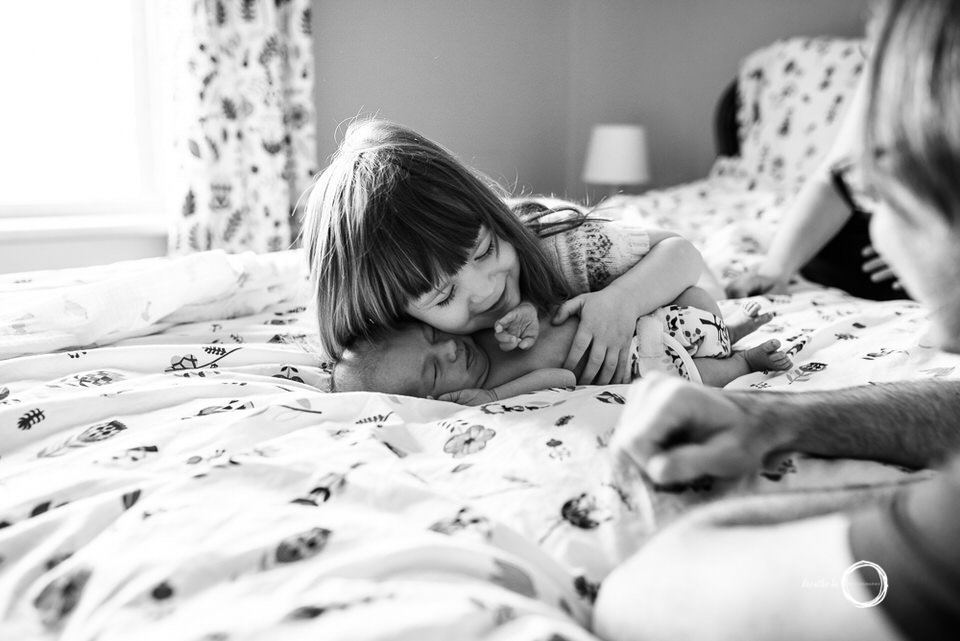 Toddler big sister cuddles with her newborn baby brother while parents watch nearby.