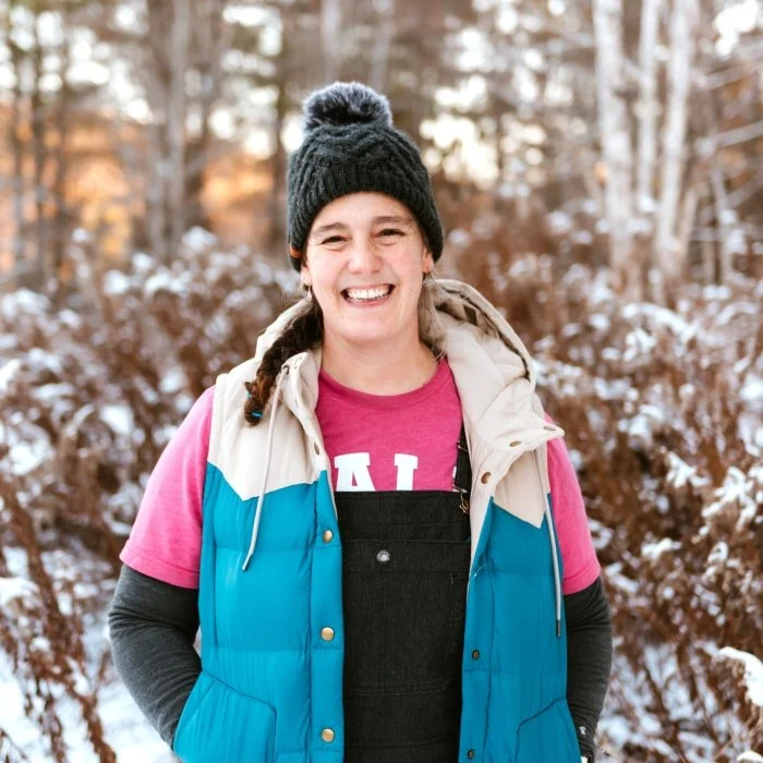 A smiling woman outdoors in a snowy winter setting, wearing a black beanie, a pink shirt, black overalls, and a blue and beige puffer vest.