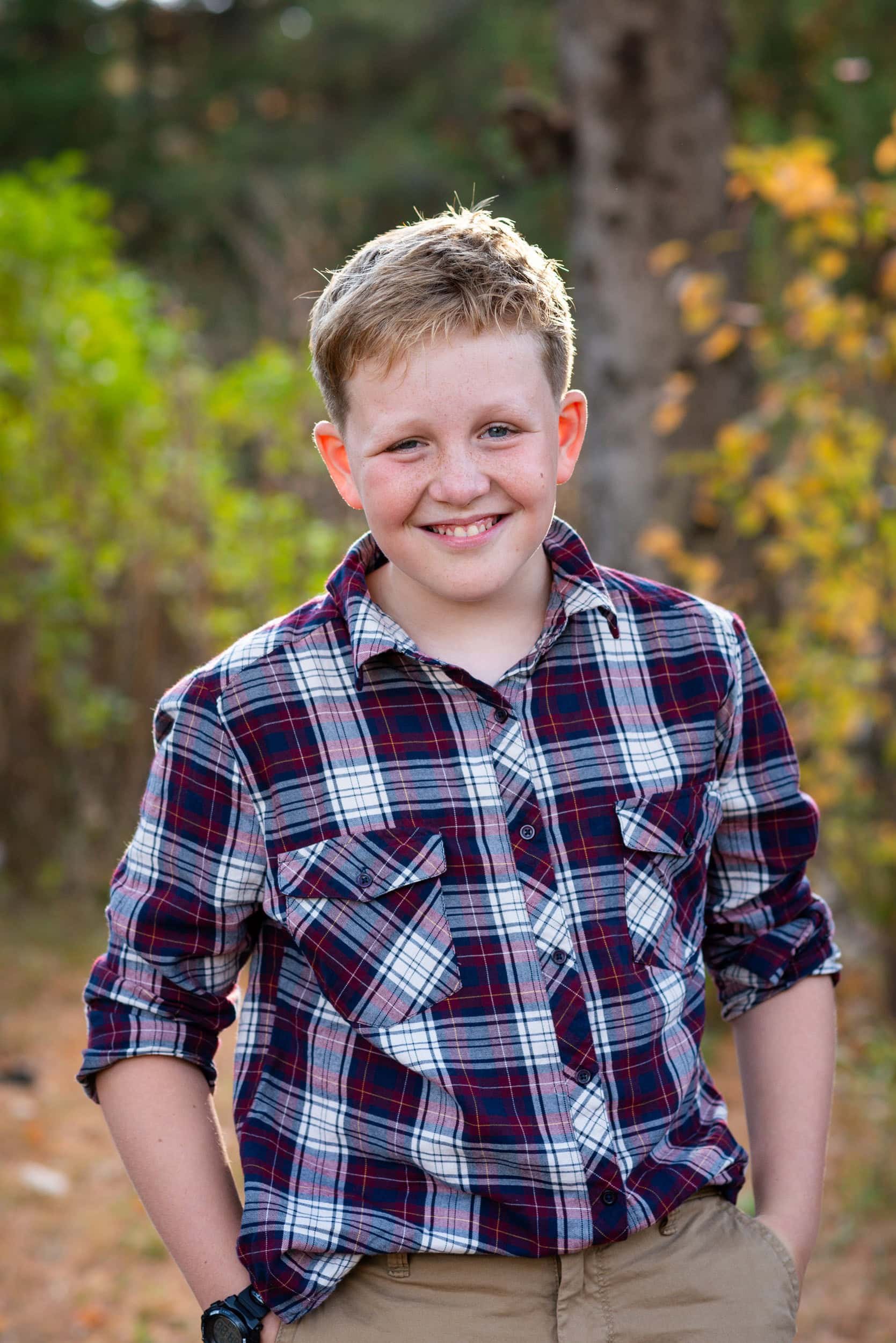 Older child with short blond hair, plaid shirt with his hands in his pockets with a warm smile in the forest.