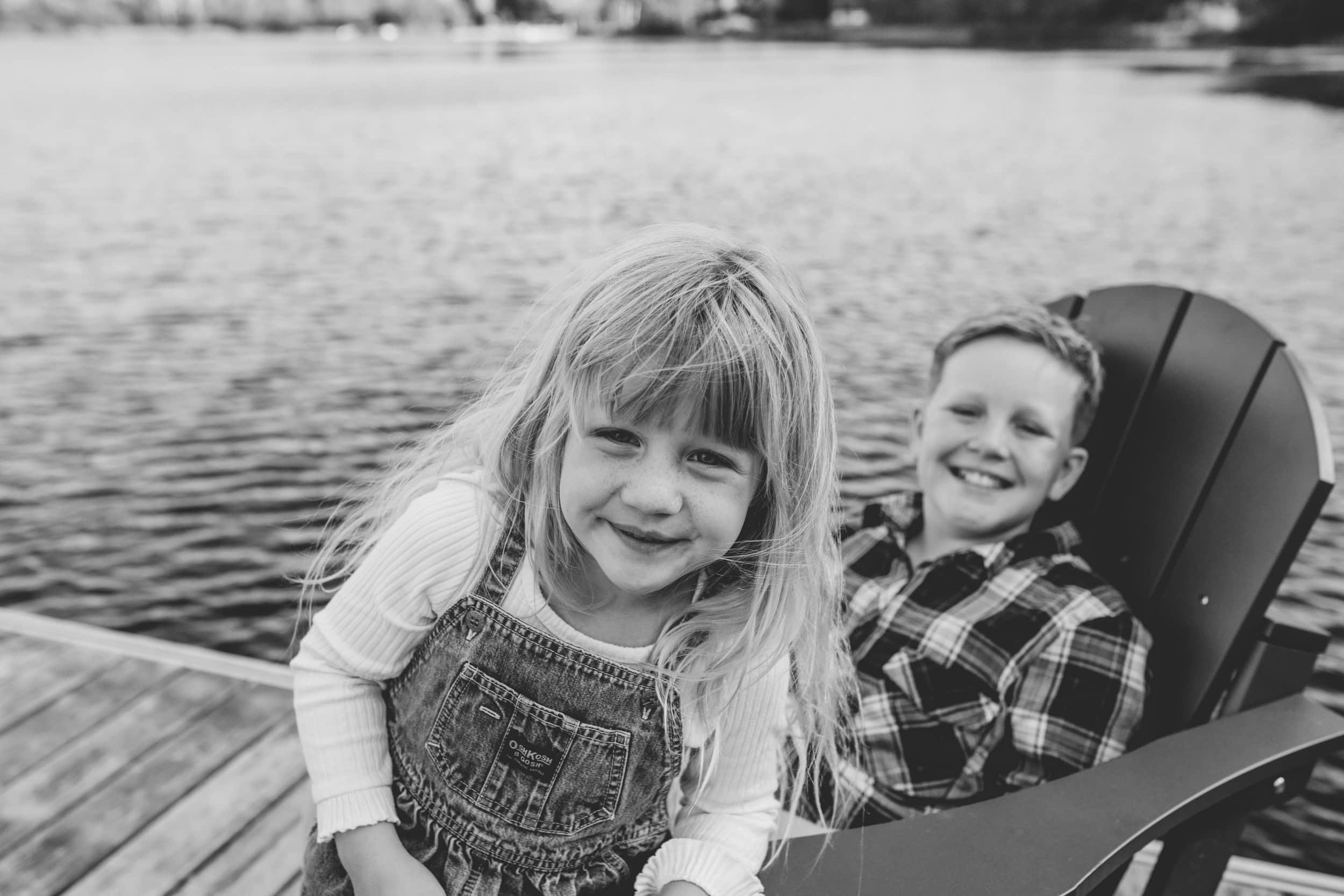 Long blond haired child with hair blowing in the lake-breeze sitting on her brother's lap on a Muskoka Chair at the lake in Val des Monts, Quebec.