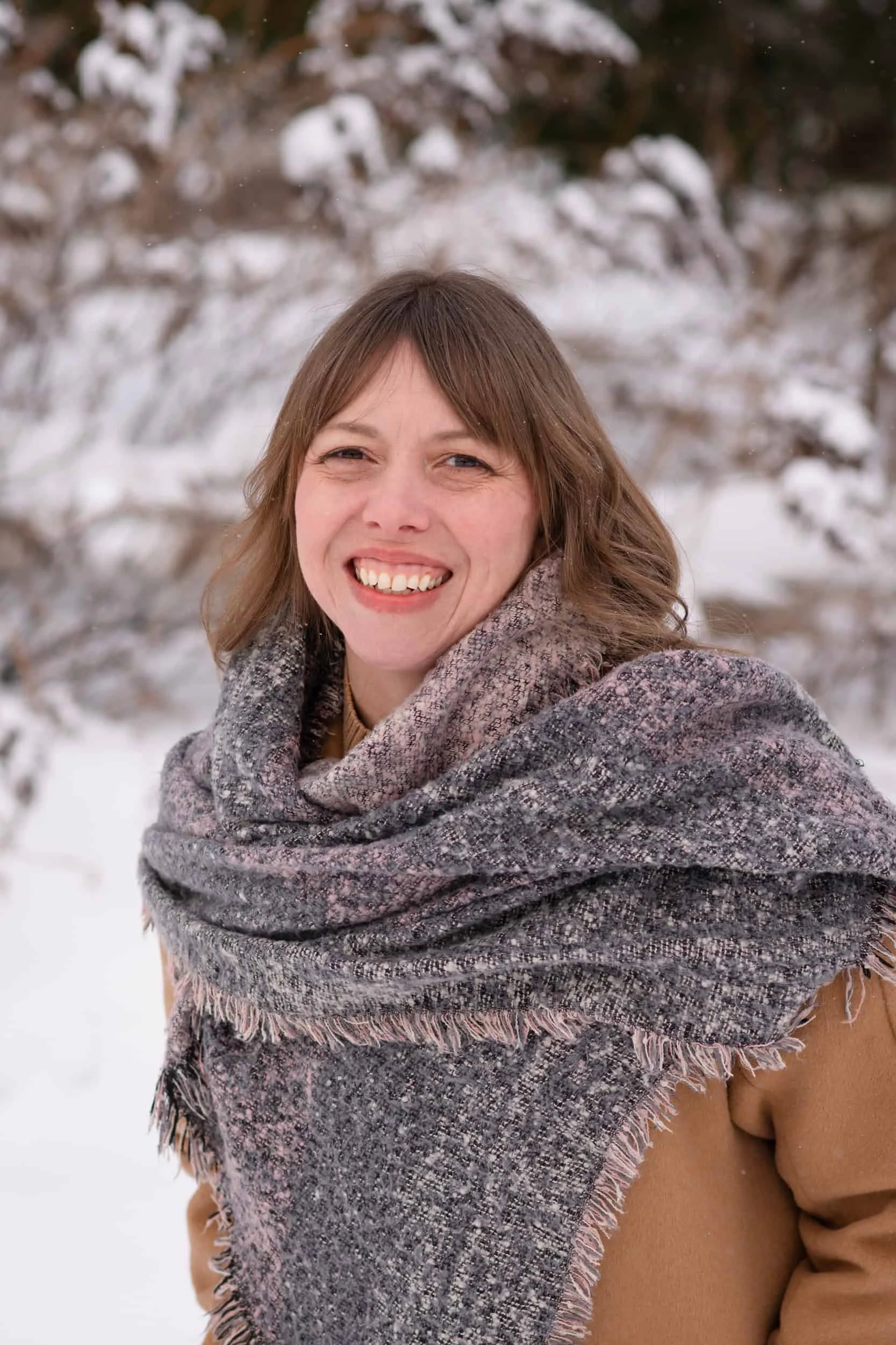 Woman with medium length brown hair standing in the snow with a pink and grey scarf over her shoulders and brown jacket.