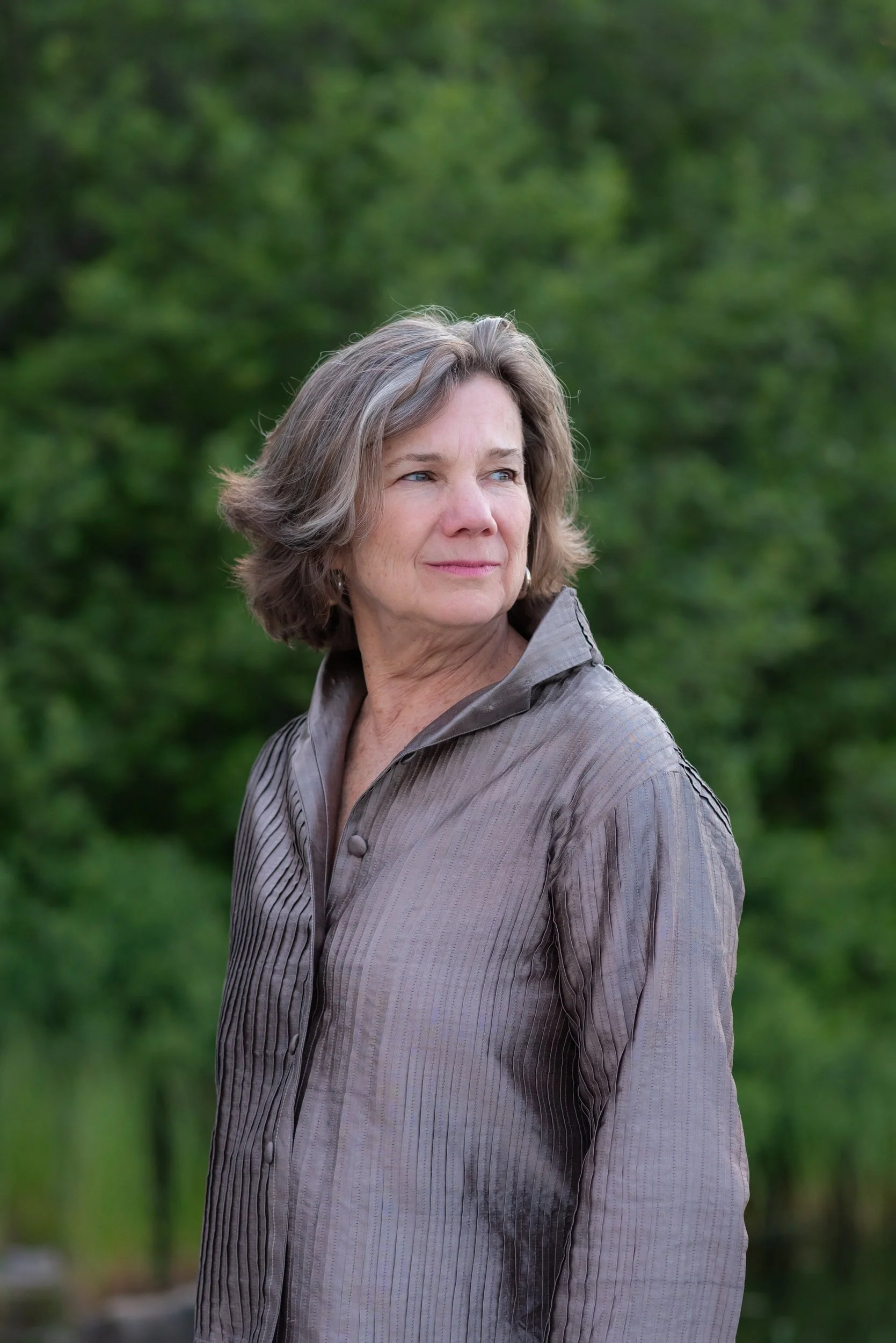 Woman standing at lake with green trees behind her and brown shirt looking out at the lake.
