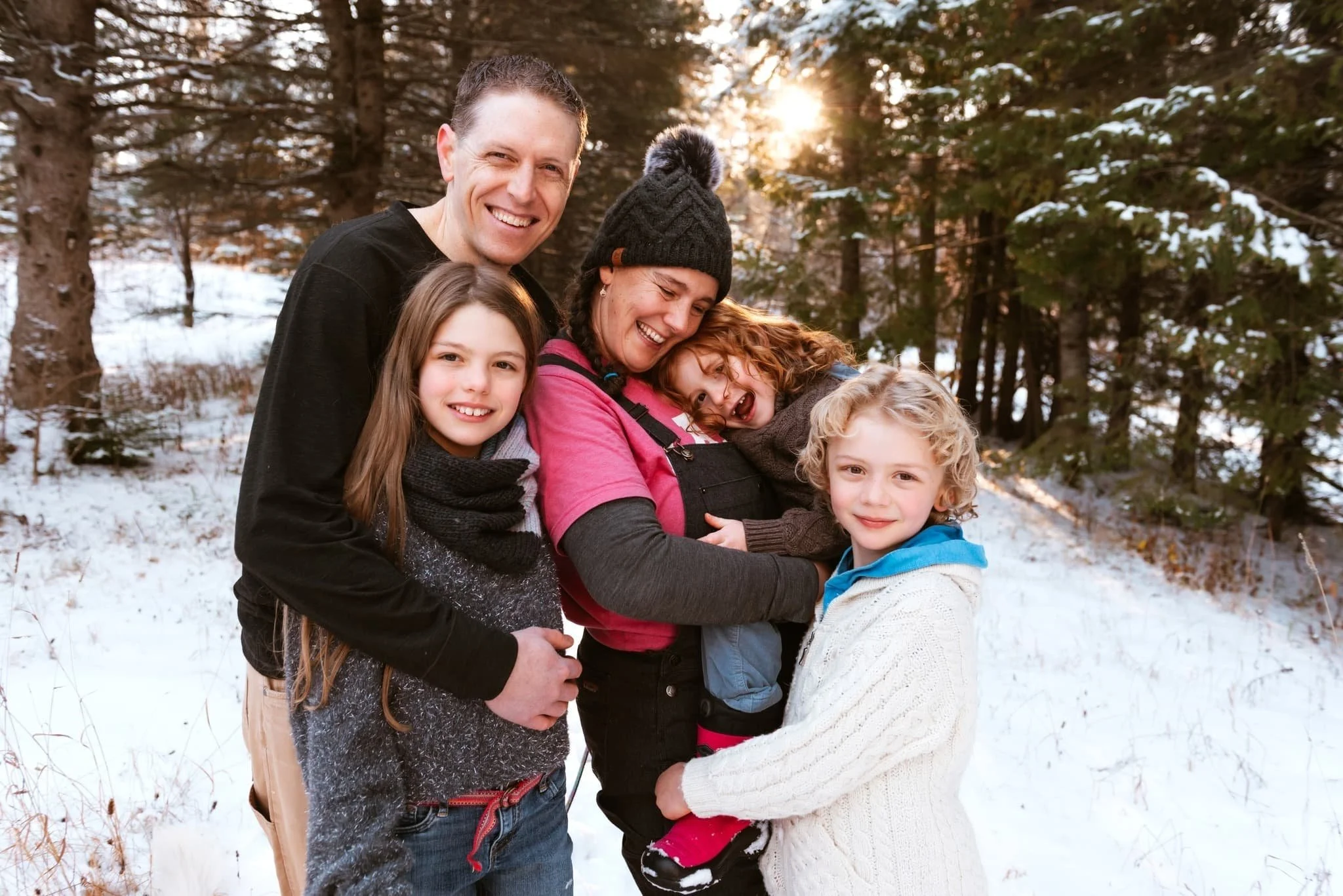 A family of  five smiling and hugging each other outdoors in a snowy forest during sunset.