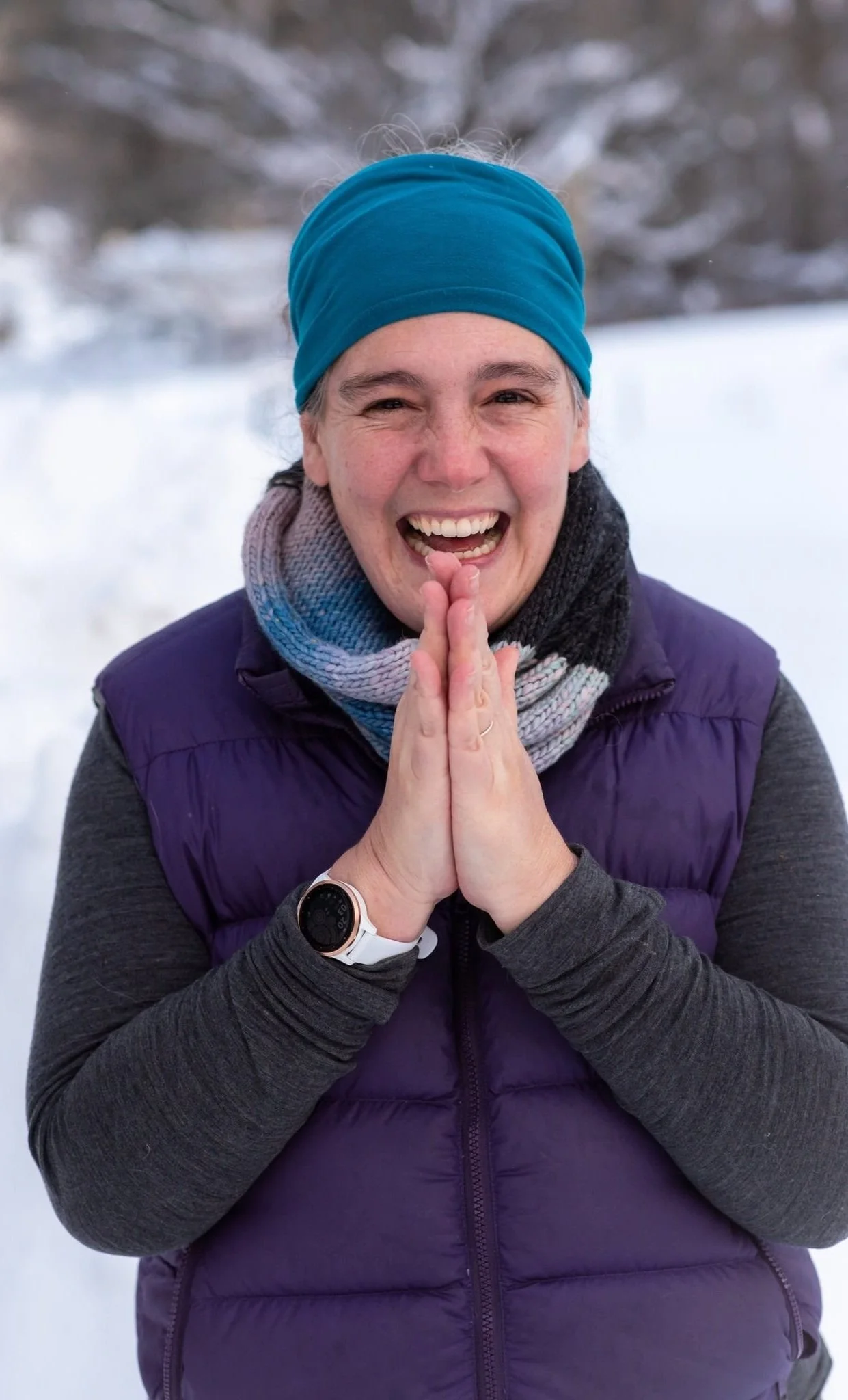 Smiling woman in winter clothing standing outdoors in snow, with hands clasped together, wearing a blue headband, purple padded vest, gray long sleeves, a multicolored scarf, and a smartwatch.