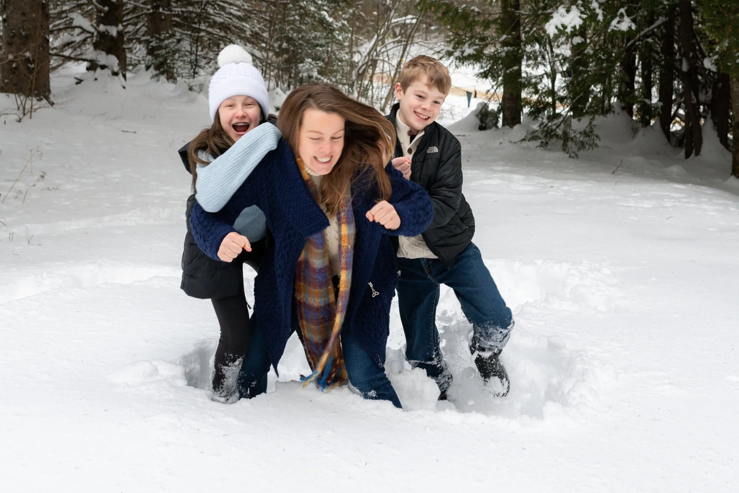 Mom and two children playing in the snow in a snowy outdoor area surrounded by trees in Val des Monts, Quebec.