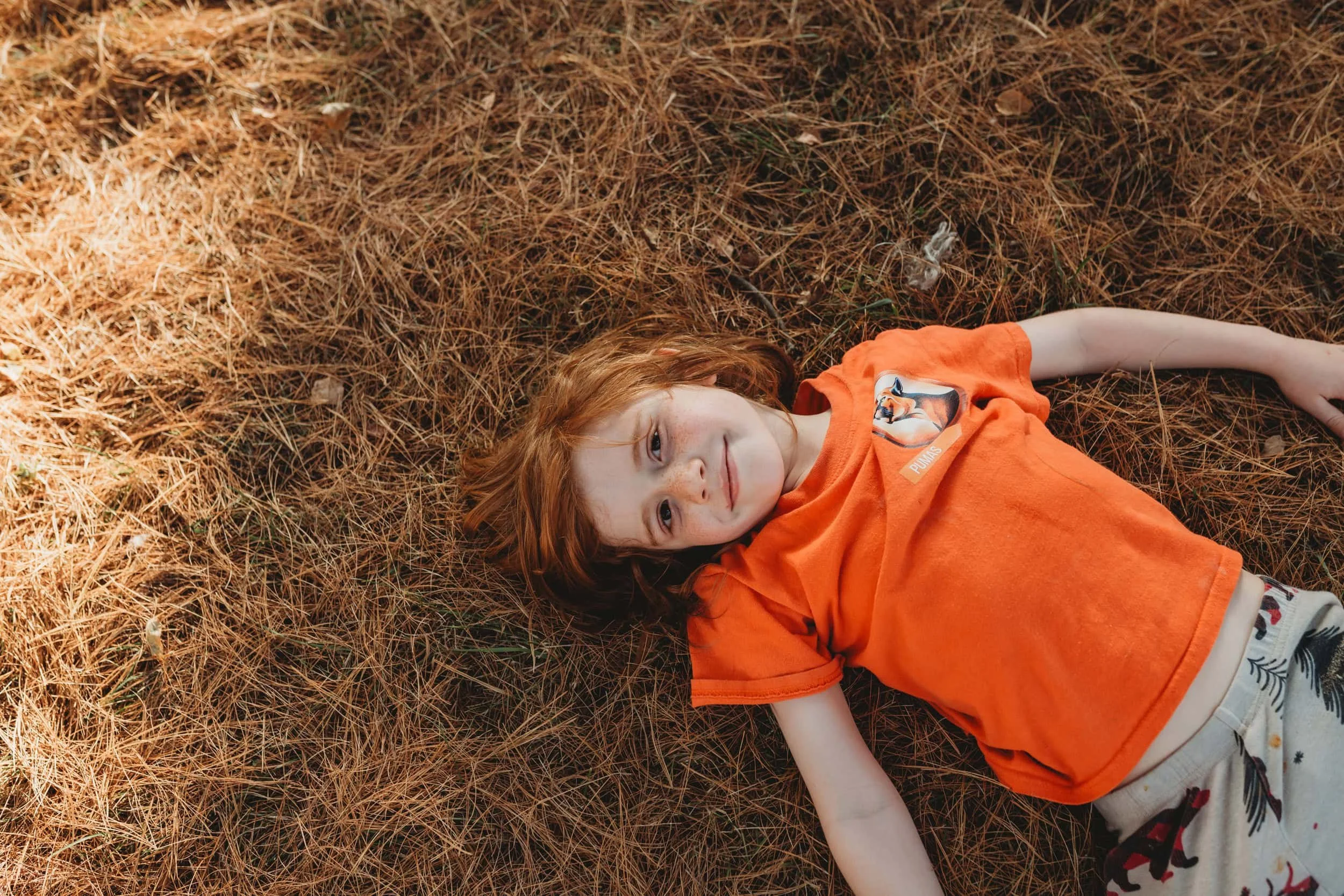 Red-headed young child laying in pine needles in the forest smiling at the camera looking very relaxed.