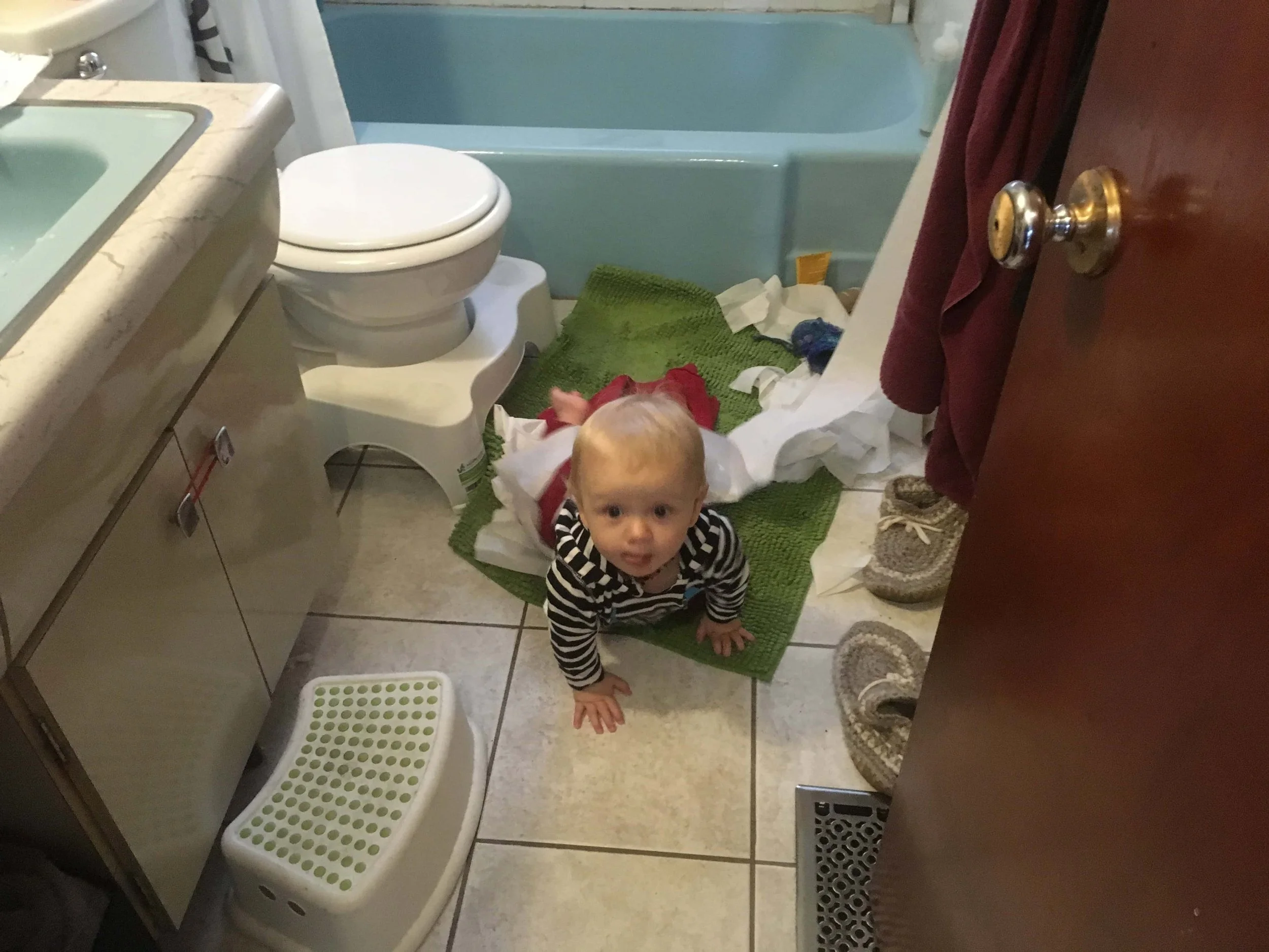 10 month old baby with toilet paper behind him and slippers beside him and cream behind him and he is blurry and crawling away from the crime scene. There is an elastic on the cabinet to keep baby out.