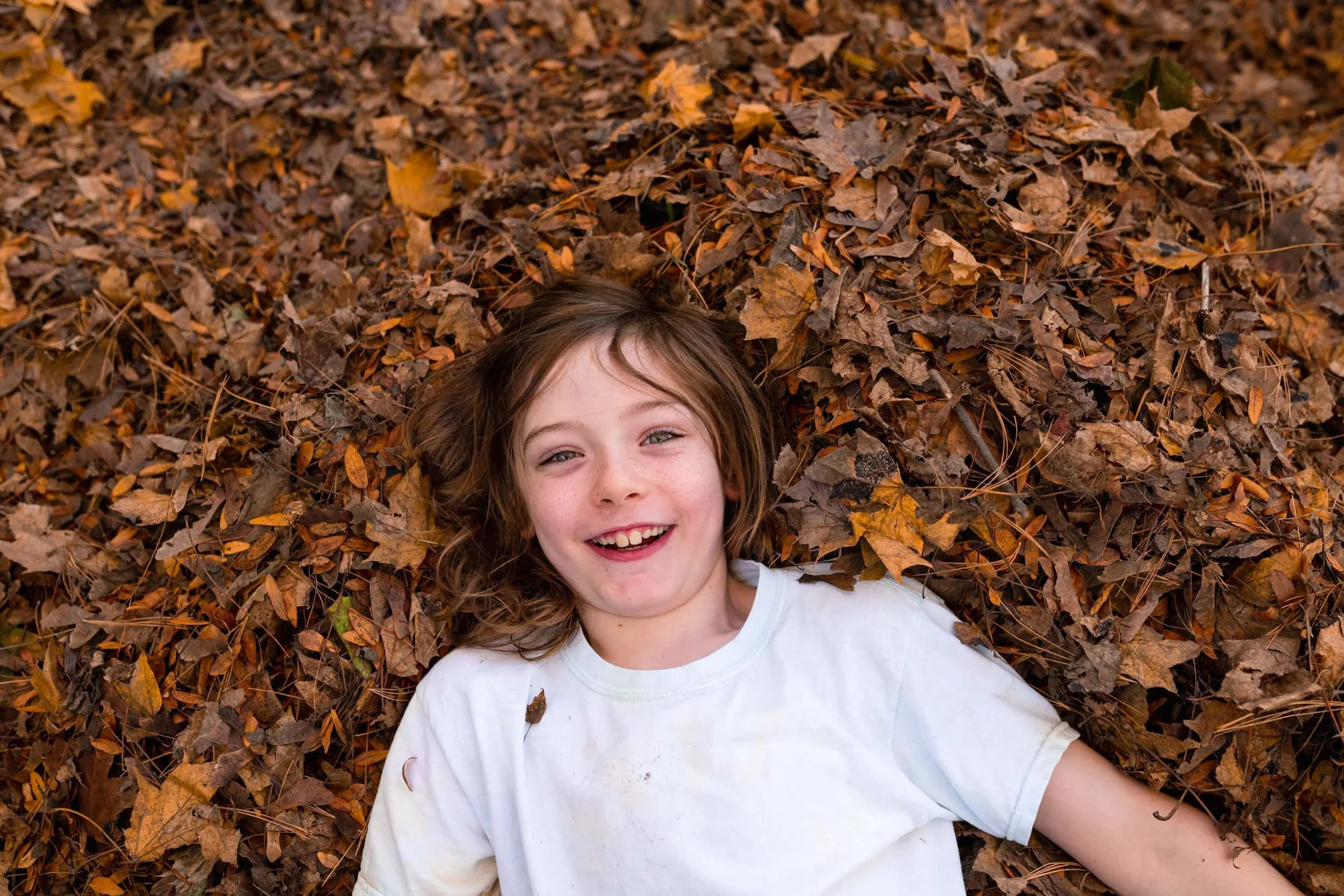 Young boy laying in fall leaves laughing wearing a white t-shirt.