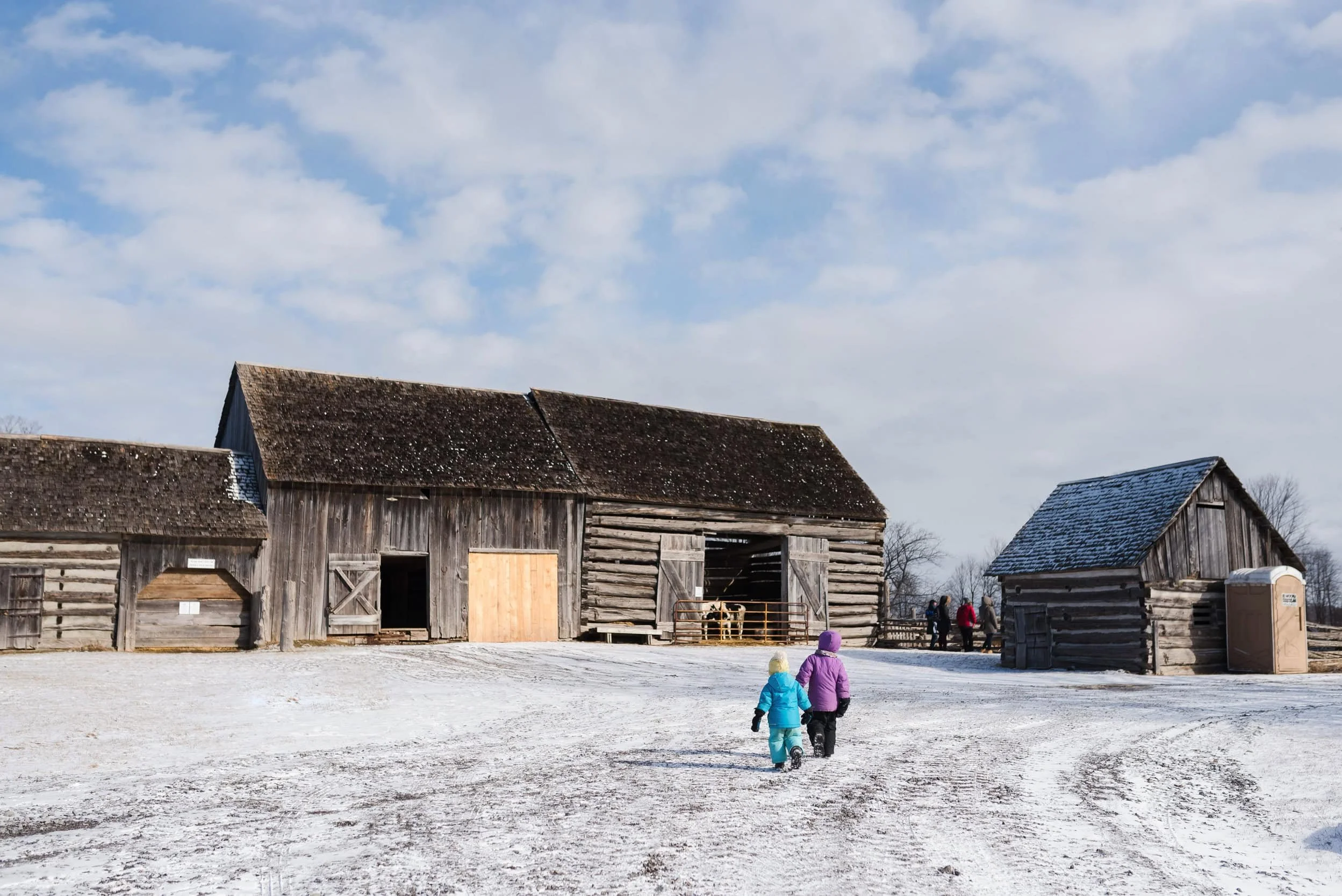 Two children walking towards old wooden barns on a snowy farm during daytime with a cloudy sky.