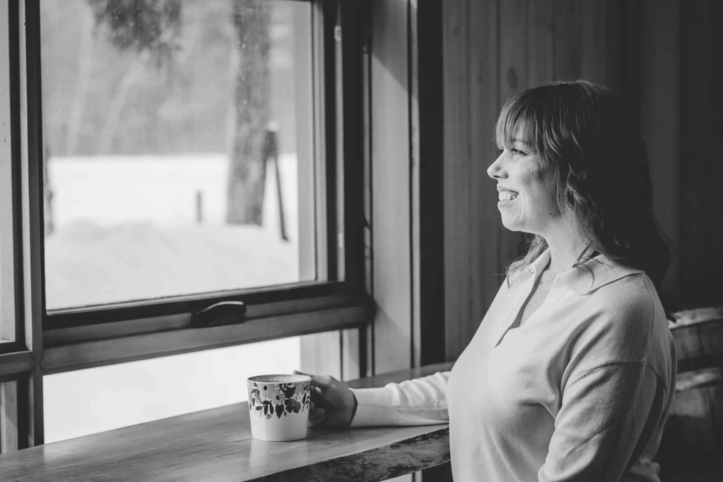 Woman drinking coffee in the window looking outside. The photo is in black and white and the woman is smiling warmly.