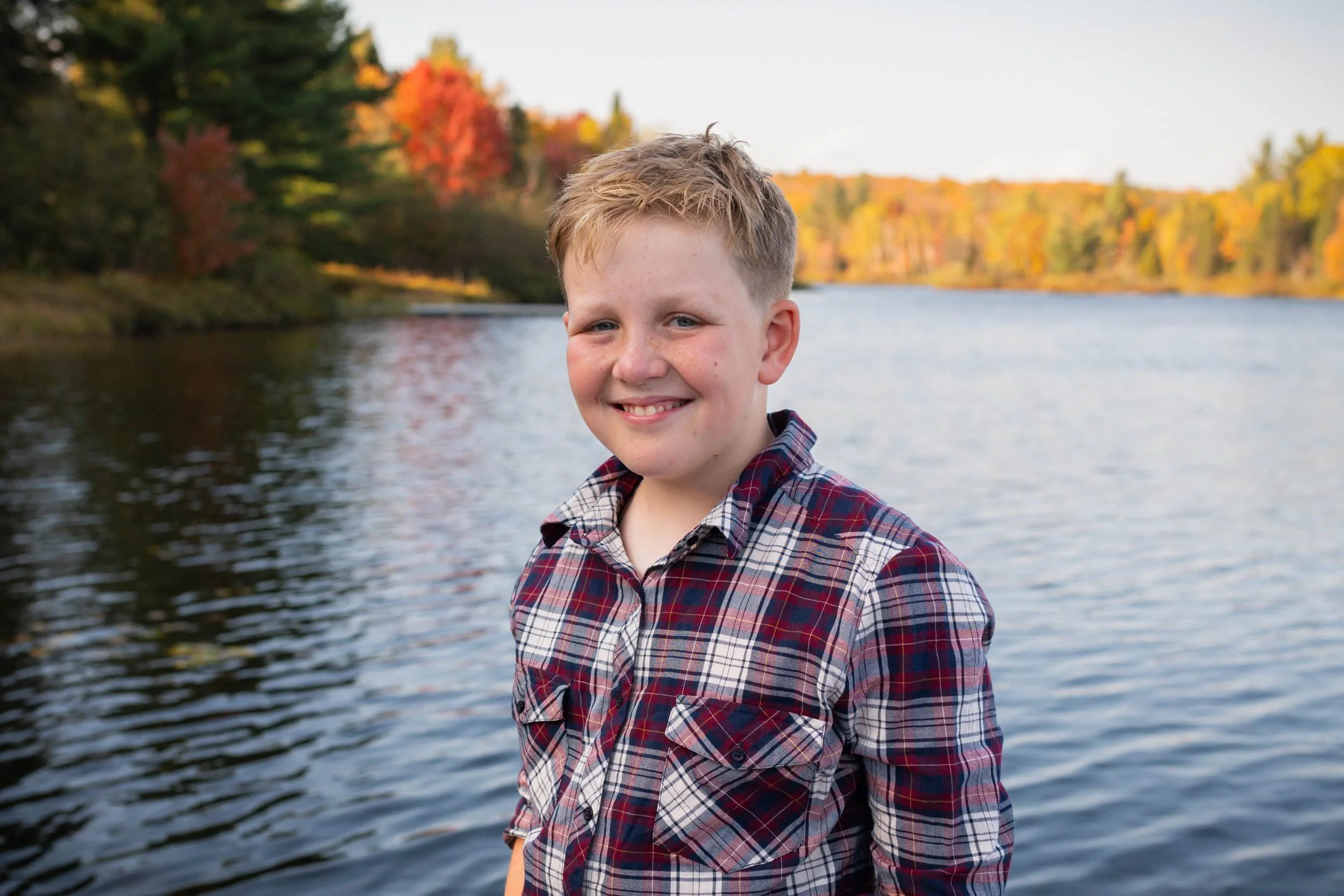 Child with blond hair and blue eyes wearing a plaid shirt at the lake at sunset in the fall.