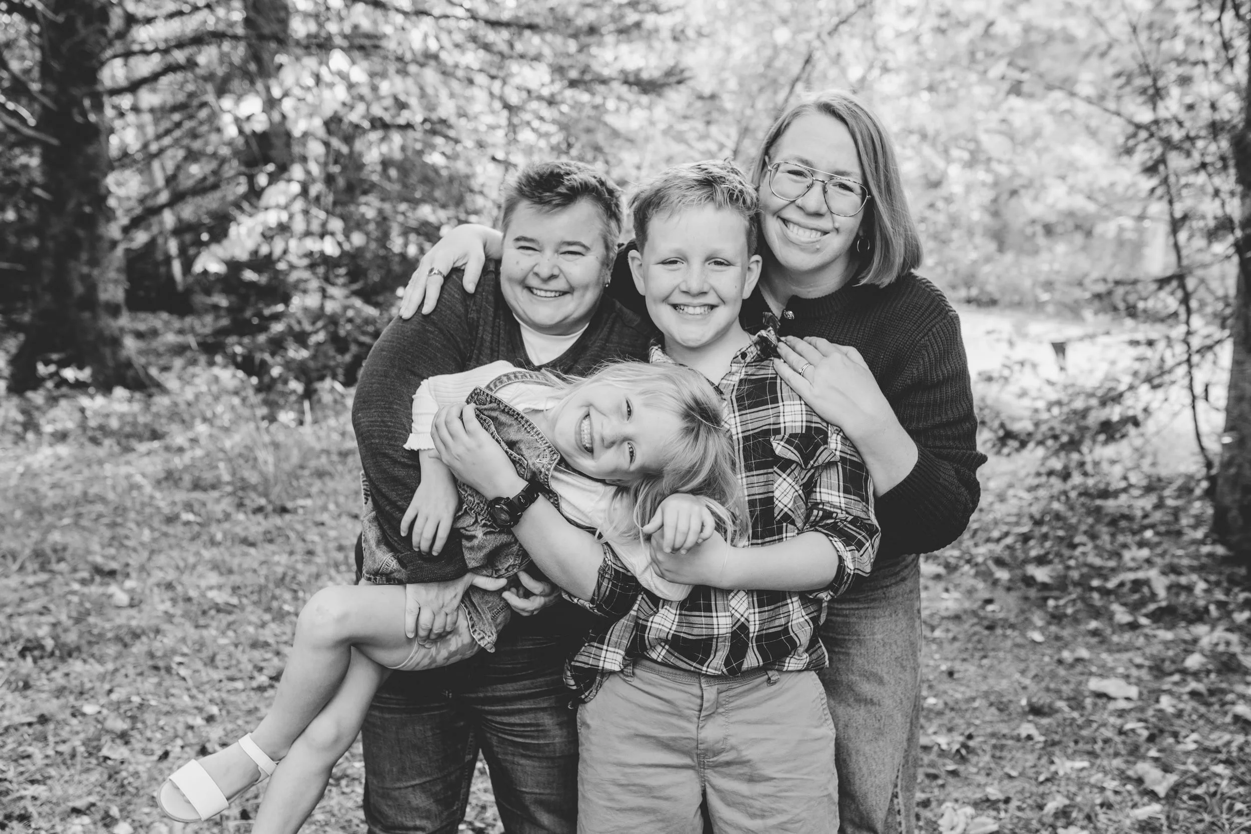 A family of four holding yougest up in the air, laughing and smiling in the forest. Very fun photo.
