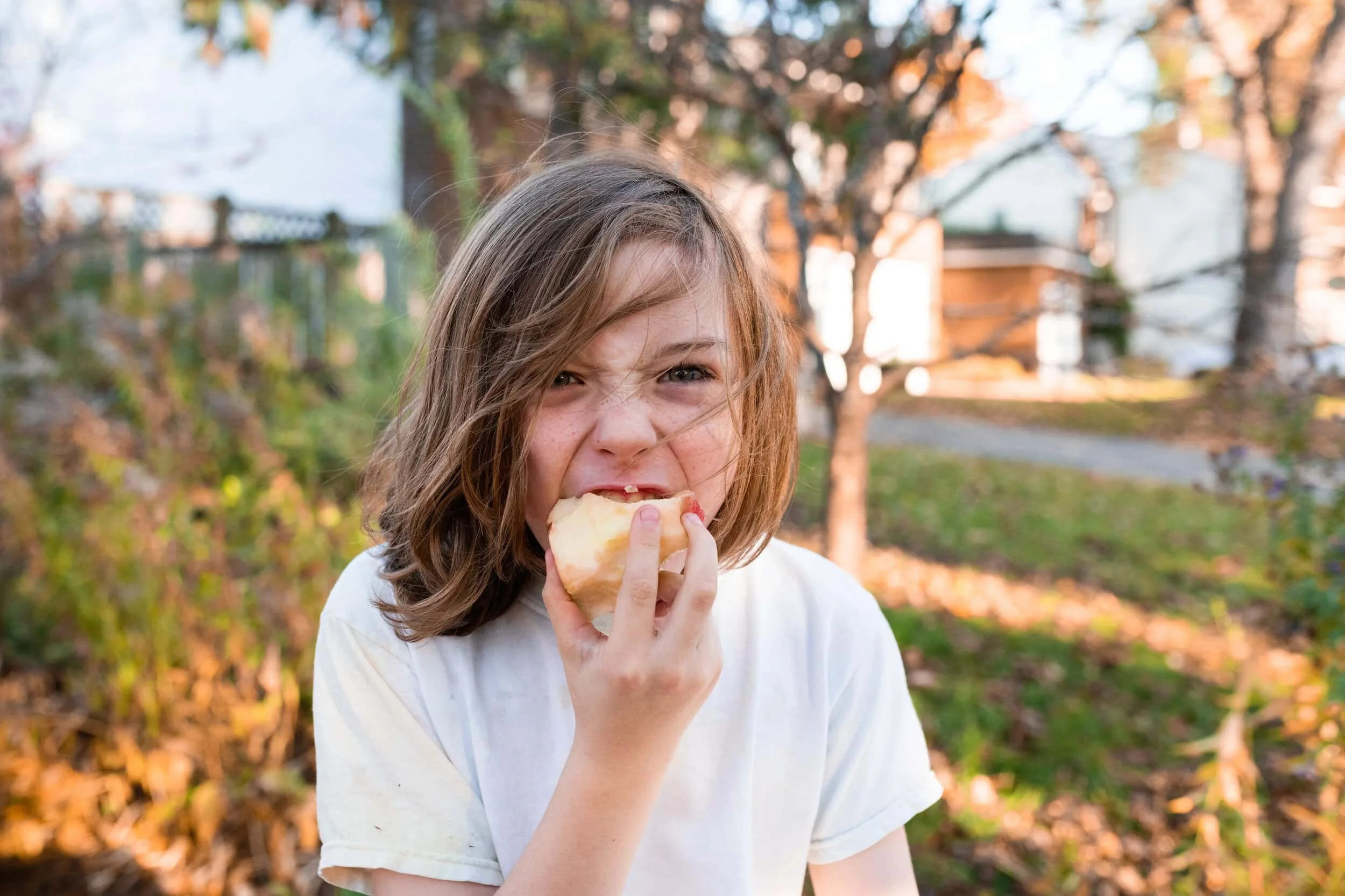 Young child with long brown hair biting into an apple looking at the camera. It is fall and the weather looks very warm.