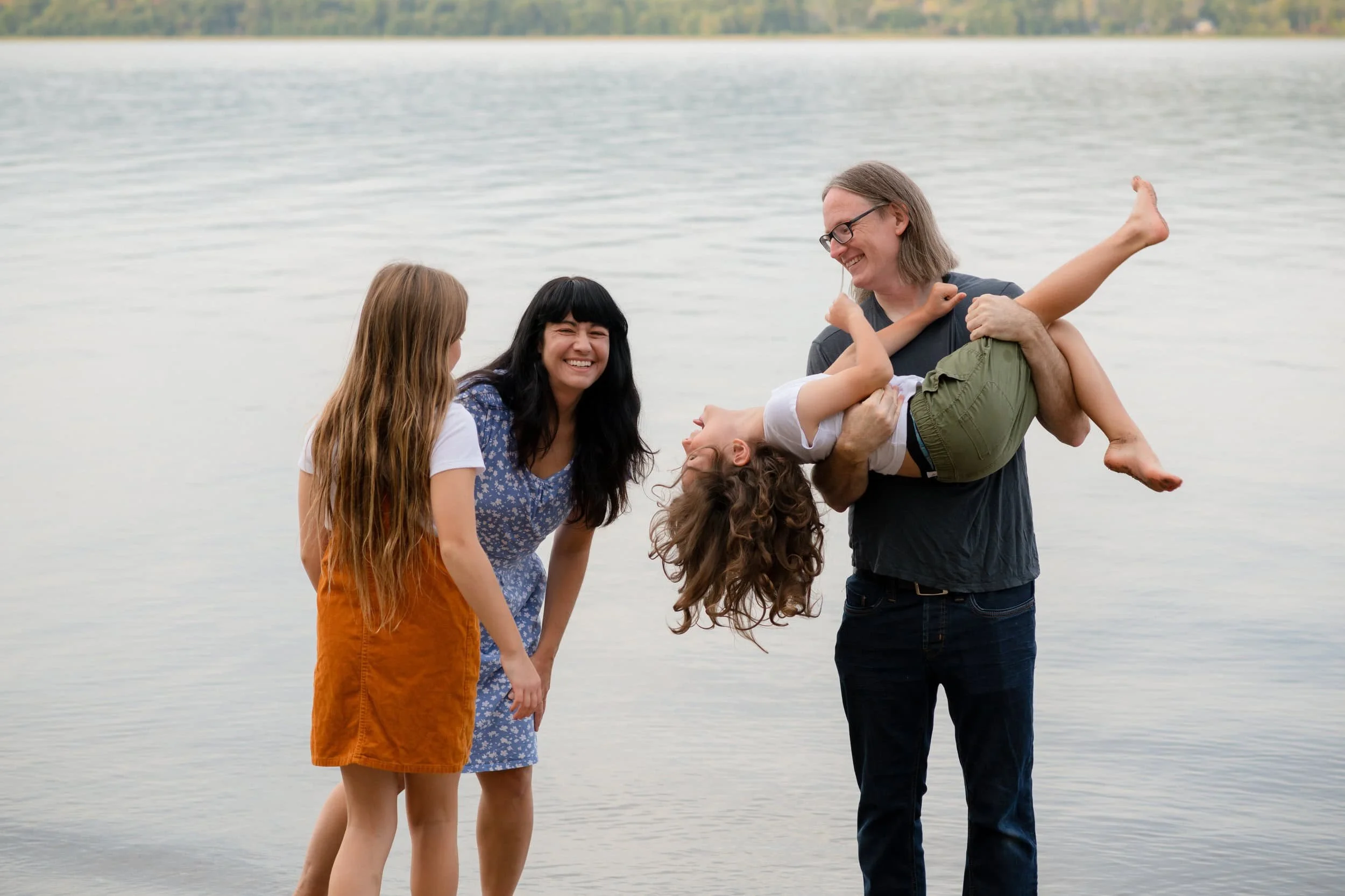 A family of four enjoying time by the water, with a man holding a young girl upside down while a woman and a girl watch and smile.