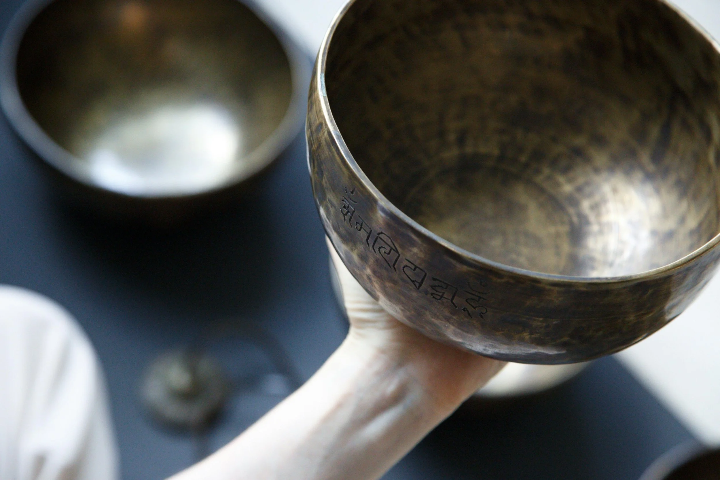 A metallic singing bowl held by a person, with a second similar bowl in the background on a dark surface.