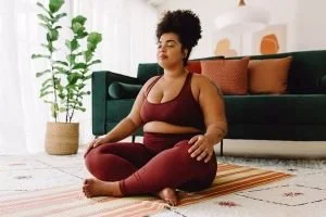 A woman practicing yoga in her living room, sitting in a cross-legged position with eyes closed, wearing a maroon sports bra and matching leggings.