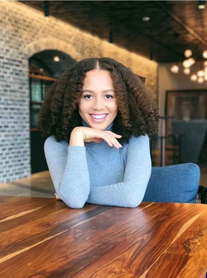 Dr. Mareena Robinson-Snowden
A young woman with curly hair smiling and resting her chin on her hand, sitting at a wooden table in a modern, stylish cafe with a brick wall and warm lighting.