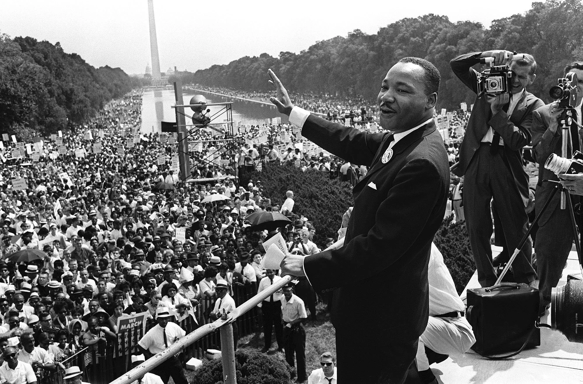A black and white photograph of Martin Luther King Jr. giving his 'I Have a Dream' speech at the March on Washington, with a large crowd of protesters holding signs and umbrellas behind him, and cameras capturing the event.