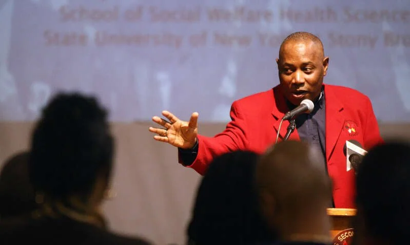 Lafred "Coach" Powell: A man in a red blazer speaking at a podium during a presentation or lecture, with an audience listening.