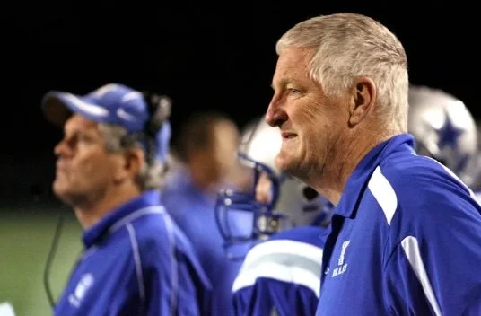Jim Place: A group of football coaches on the sidelines during a game, wearing blue jackets and helmets.