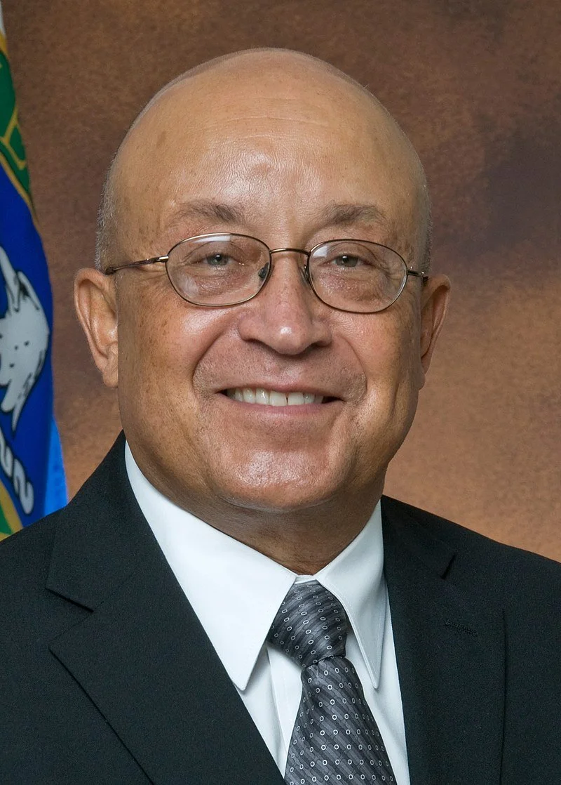 DR. WARREN F. "PETE" MILLER JR.
A smiling man in a suit, wearing glasses, with a flag partially visible in the background.