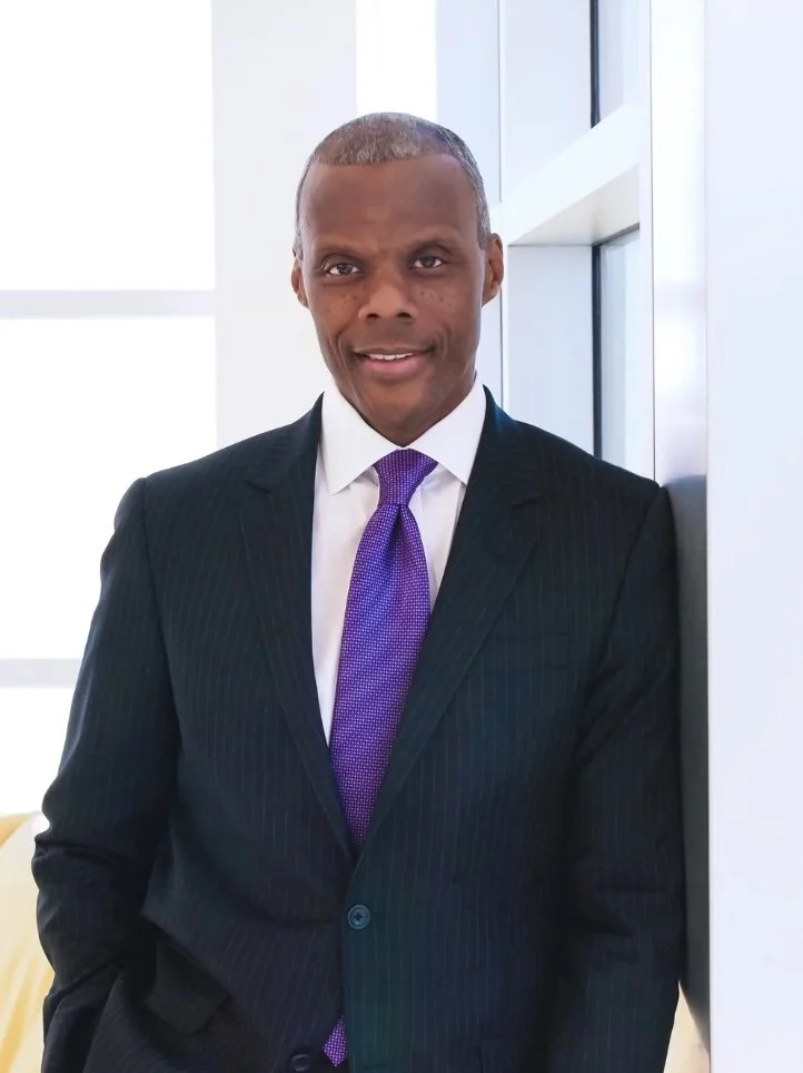 JC Watts: A man in a dark pinstripe suit with a purple tie standing in an office, leaning against a wall.
