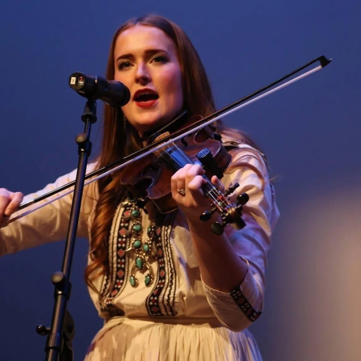 Shelby Murdock: IFAAA Performer A woman with long brown hair singing into a microphone while playing a violin on stage, wearing a beige embroidered dress.