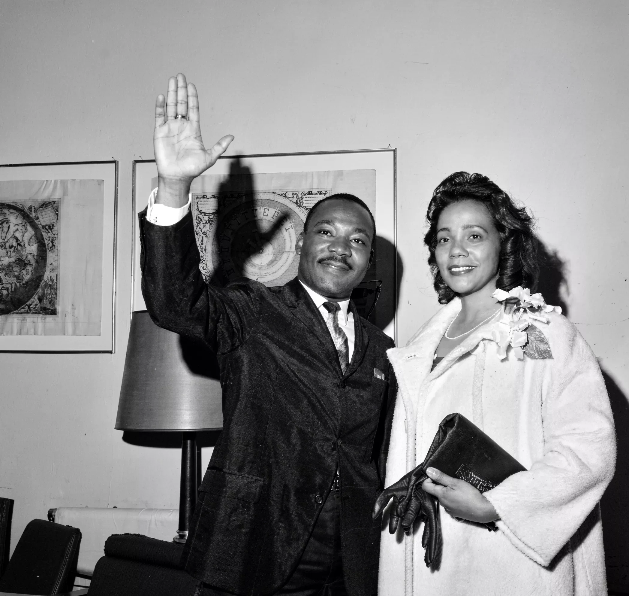 A black and white photograph of a man and a woman indoors. The man, wearing a suit, is waving with his right hand and has a slight smile. The woman, dressed in a coat with floral embellishments and holding gloves and a clutch purse, is standing beside him with a smile. There are framed artworks, a table lamp, and chairs in the background.