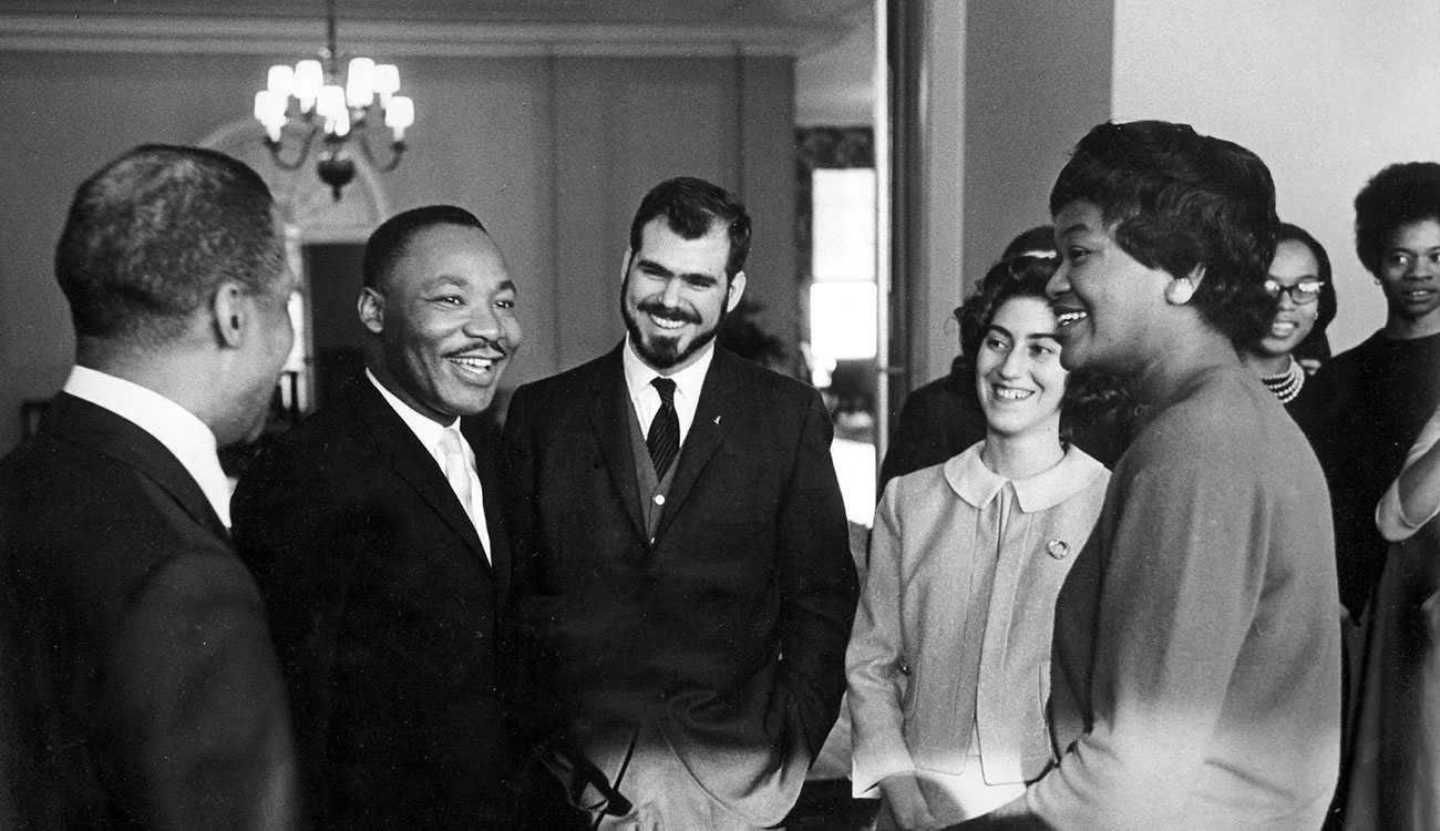 Black and white photo of a group of people, including Martin Luther King Jr. (second from left), smiling and engaging in conversation indoors with a chandelier visible on the ceiling.